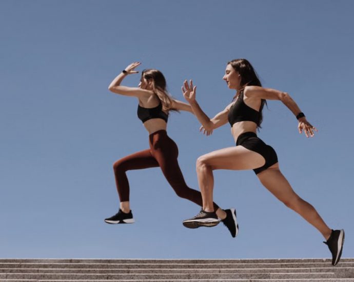 Two women in active wear leap energetically on stairs against a clear blue sky.