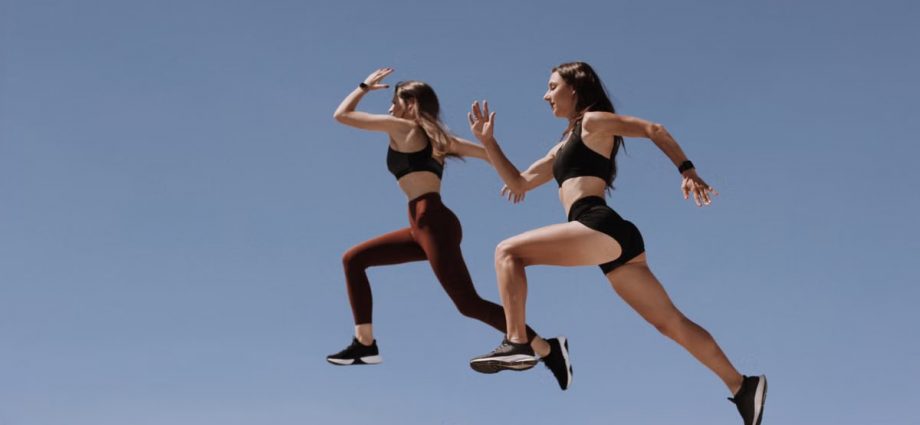 Two women in active wear leap energetically on stairs against a clear blue sky.