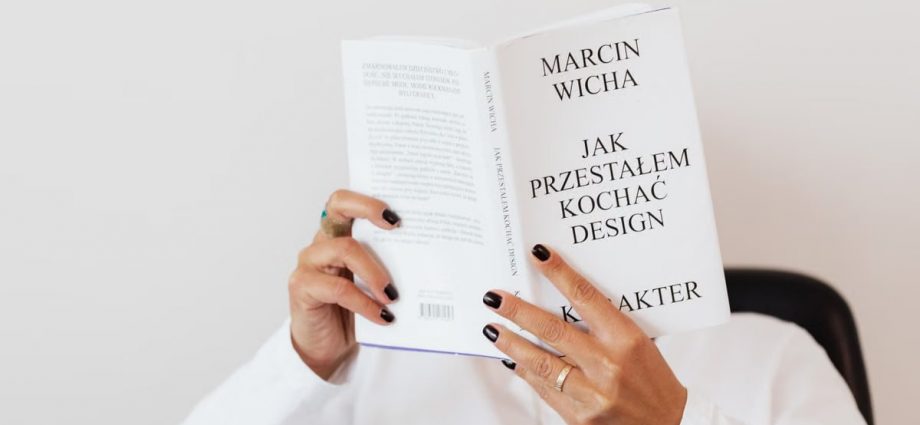 Anonymous female in white shirt with gold chain on neck reading interesting book covering face while sitting on black chair